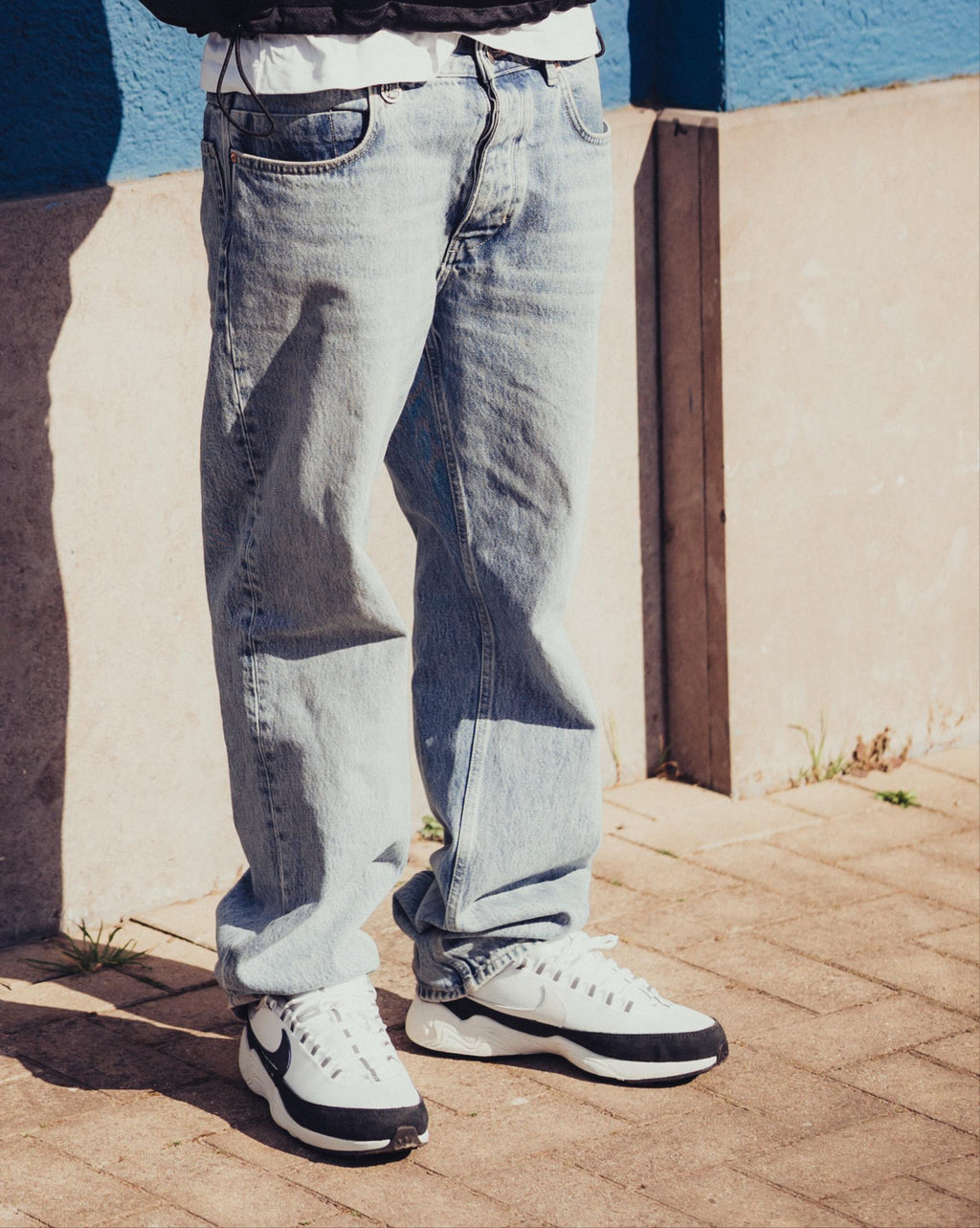 Person wearing a patterned jacket, jeans, and white sneakers against a blue wall.